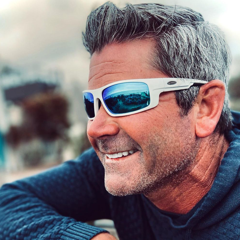 Scruffy man in sweater with salt-and-pepper gray hair wearing Sea Striker Finatic polarized sunglasses in white frame with blue mirrored lenses