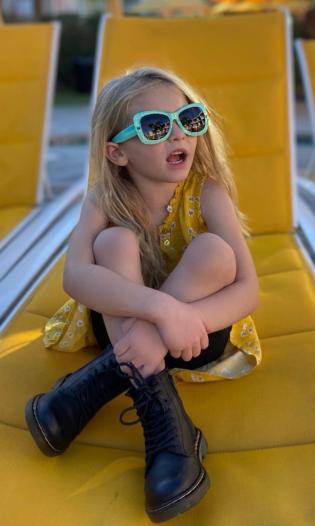 Young girl wearing Just A Shade Smaller Smiley sunglasses and boots sitting on yellow lounge chair